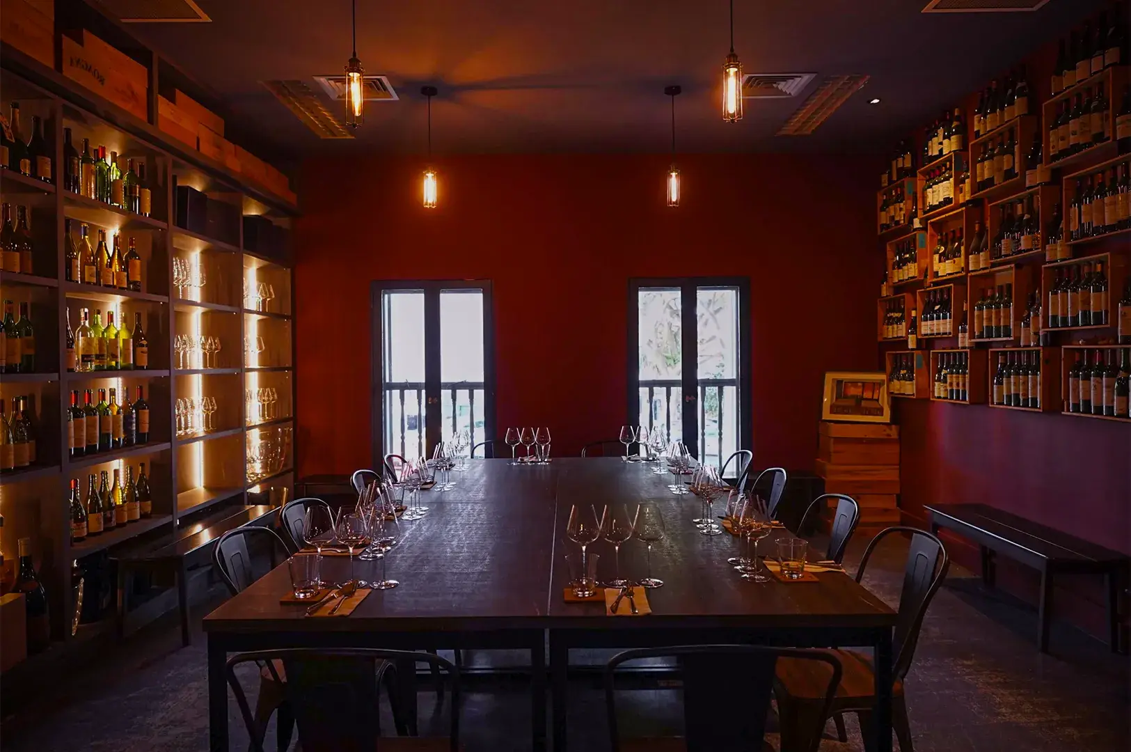 Dimly lit wine cellar dining room with red walls, wooden table set for 12, surrounded by shelves filled with wine bottles. Warm, cozy atmosphere.