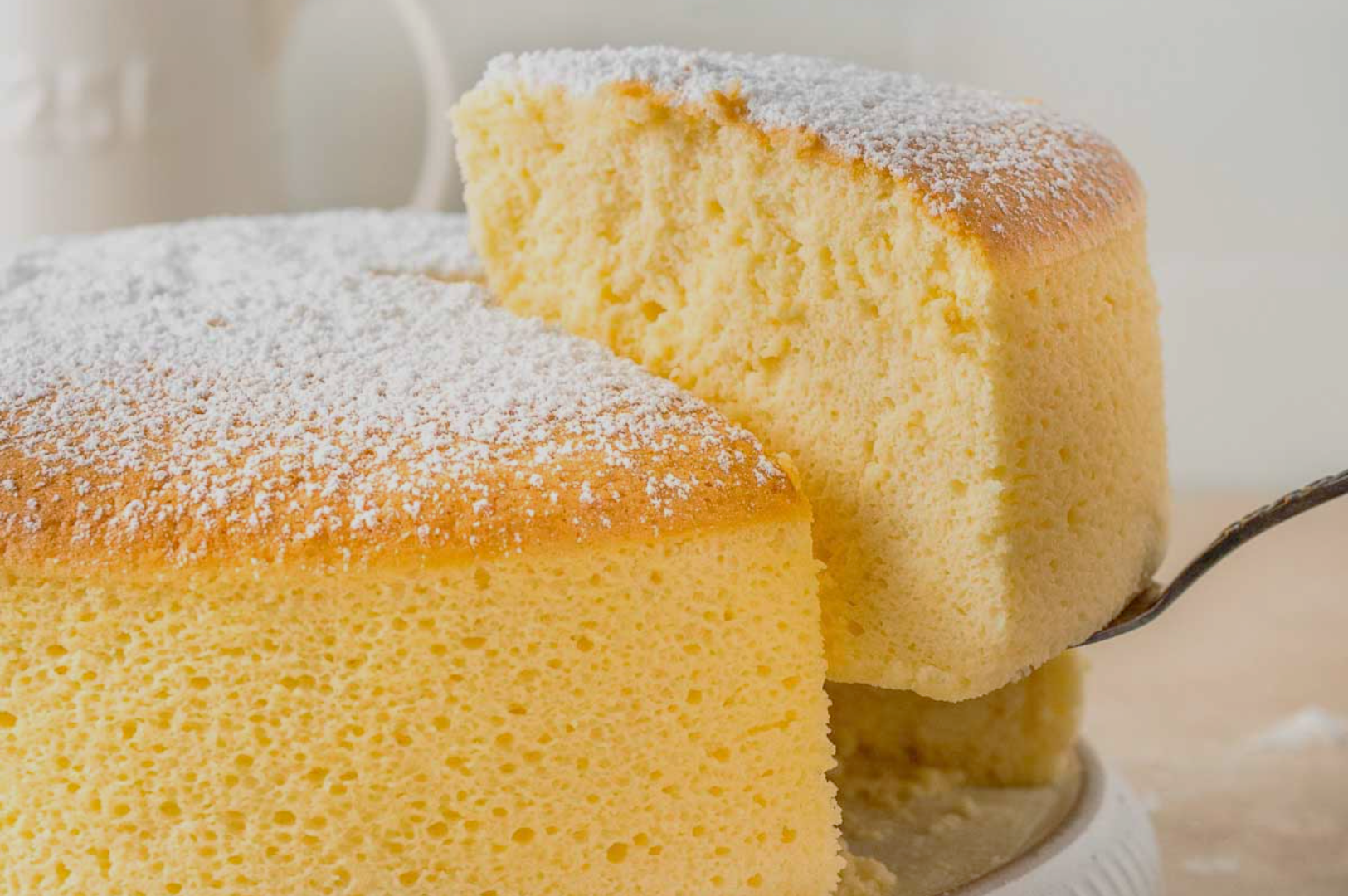 A fluffy, golden sponge cake dusted with powdered sugar has a slice lifted by a spatula. The cake's texture appears soft and airy, evoking a light, delicate feel.