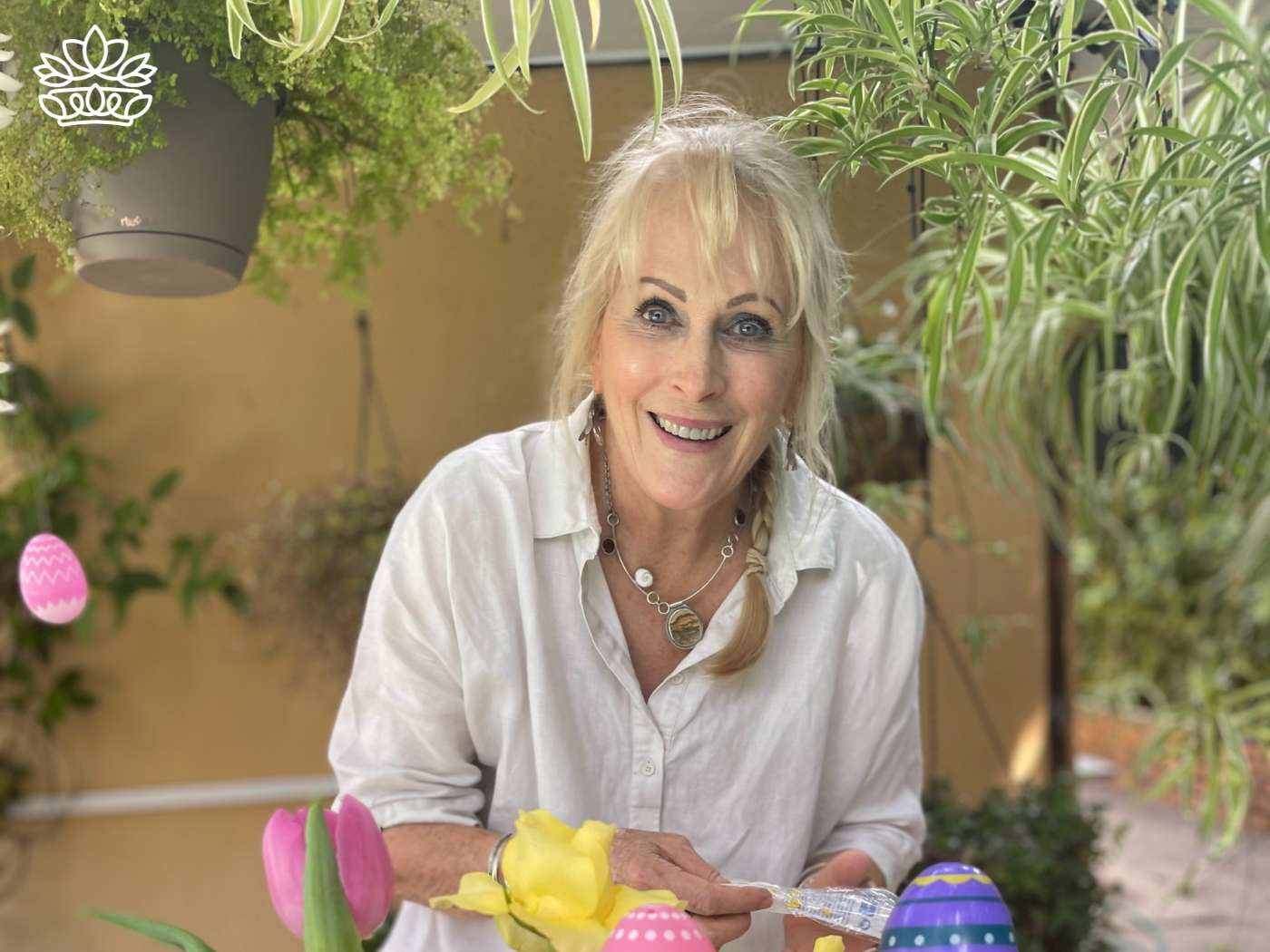 Smiling woman creating a traditional Easter basket with hot glue, surrounded by colorful decorations, showcasing the larger Easter basket variety from the Easter Collection by Fabulous Flowers and Gifts.