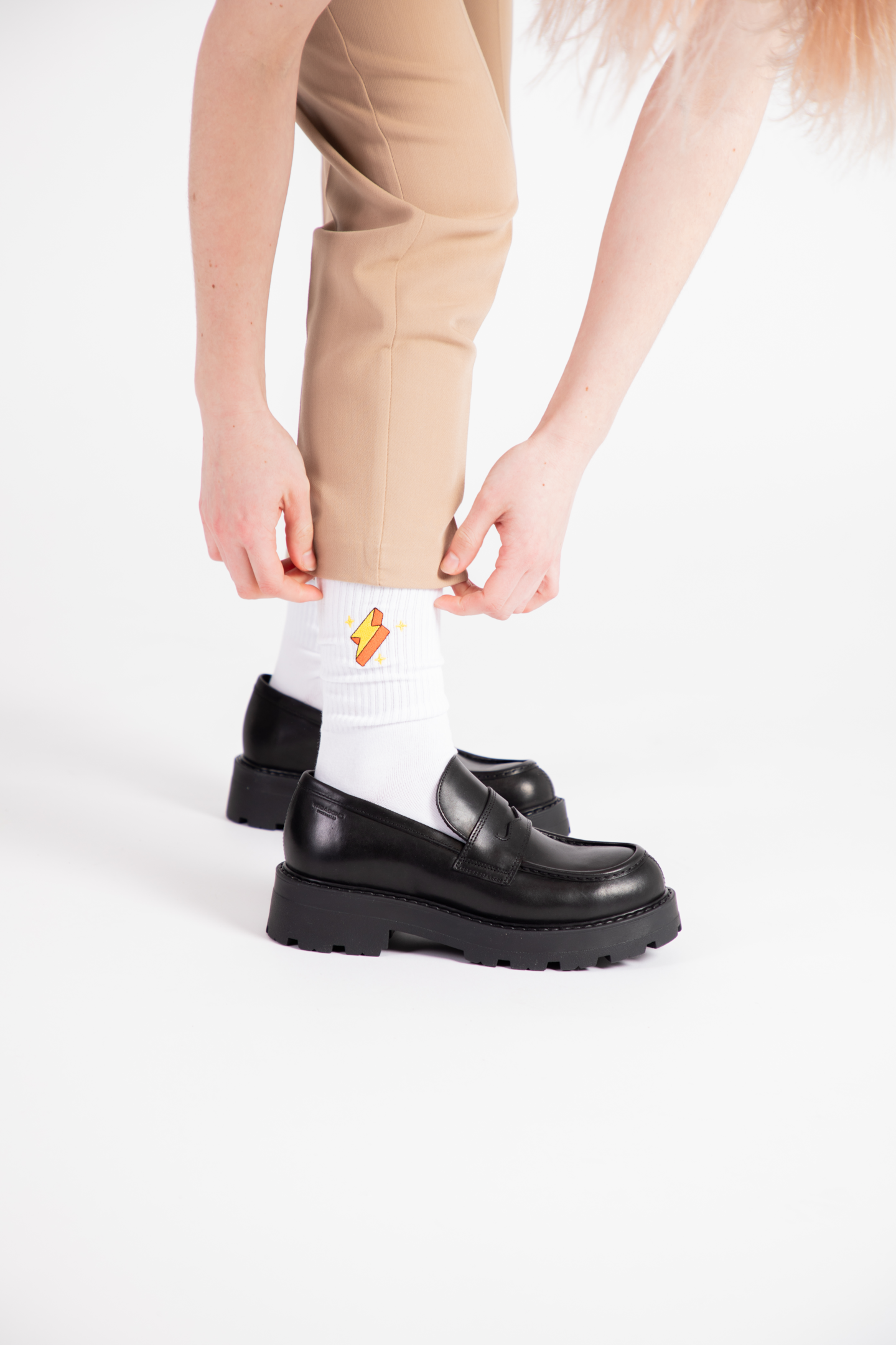 A female model wears black leather loafers with a pair of white crew socks with a lightening bolt embroidered.