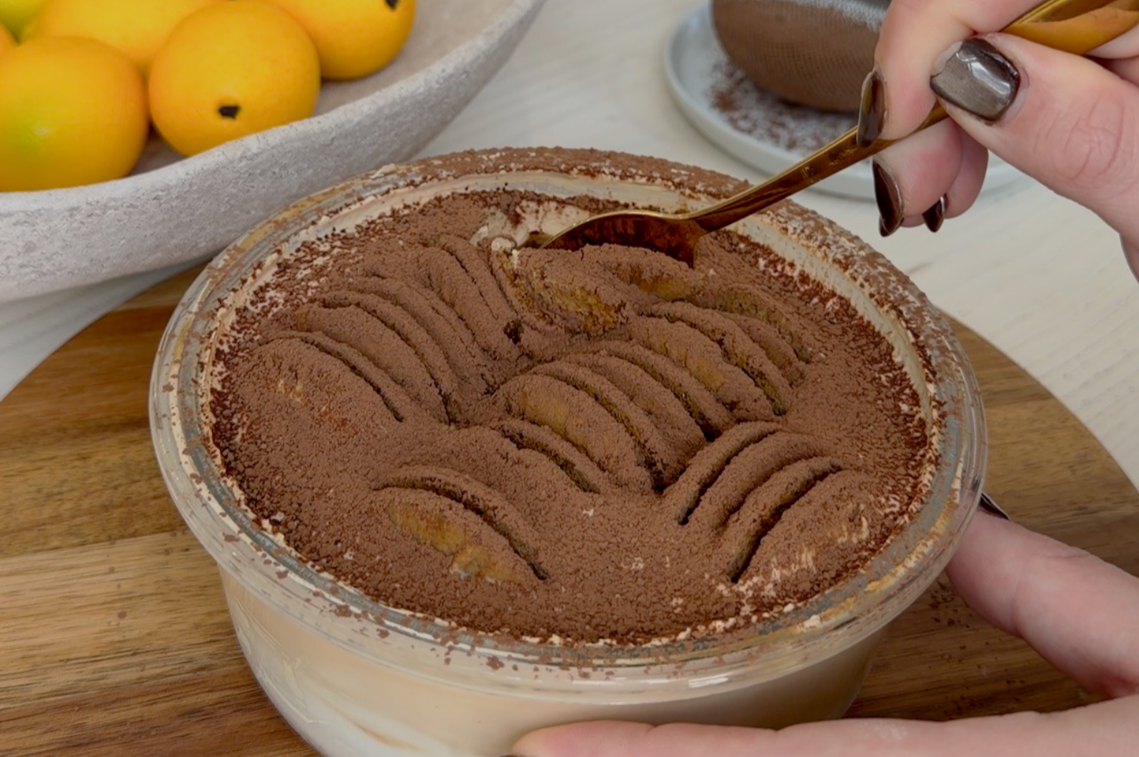 A person uses a spoon to scoop a layer of cocoa-coated tiramisu from a glass bowl. Nearby, a bowl of bright citrus fruits adds a fresh contrast.