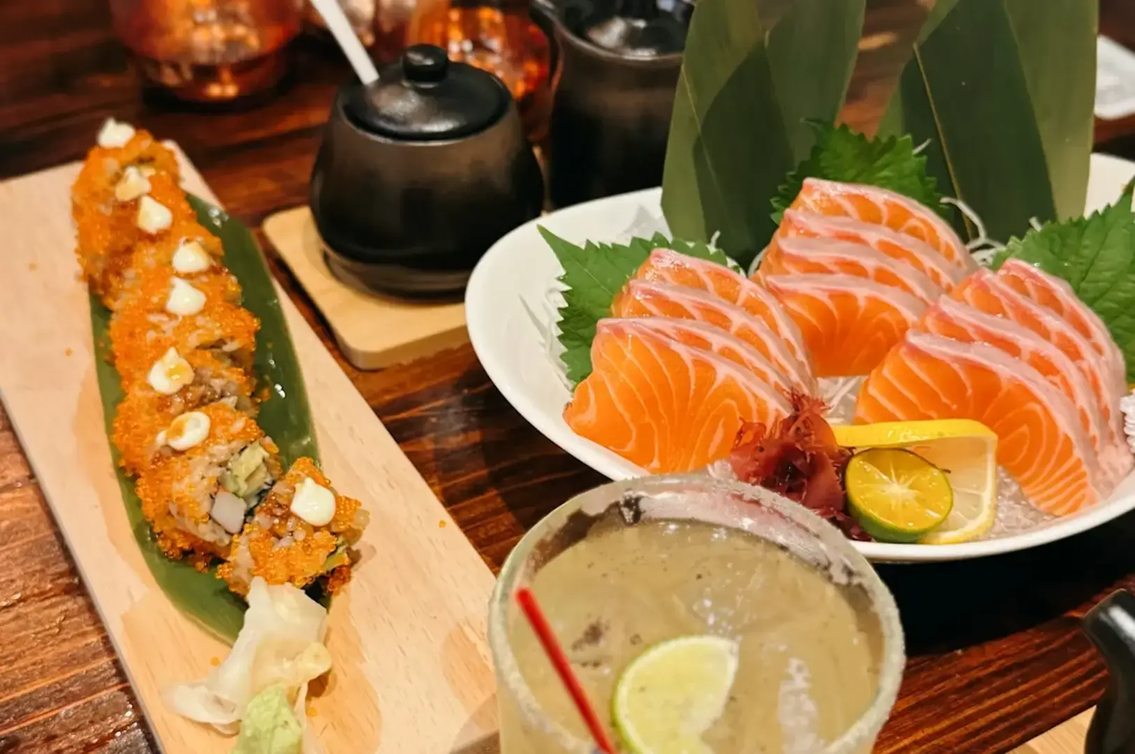 Sliced salmon sashimi on a white plate beside a sushi roll on a wooden board, with lime, pickled ginger, dipping sauce, and a drink on the table.