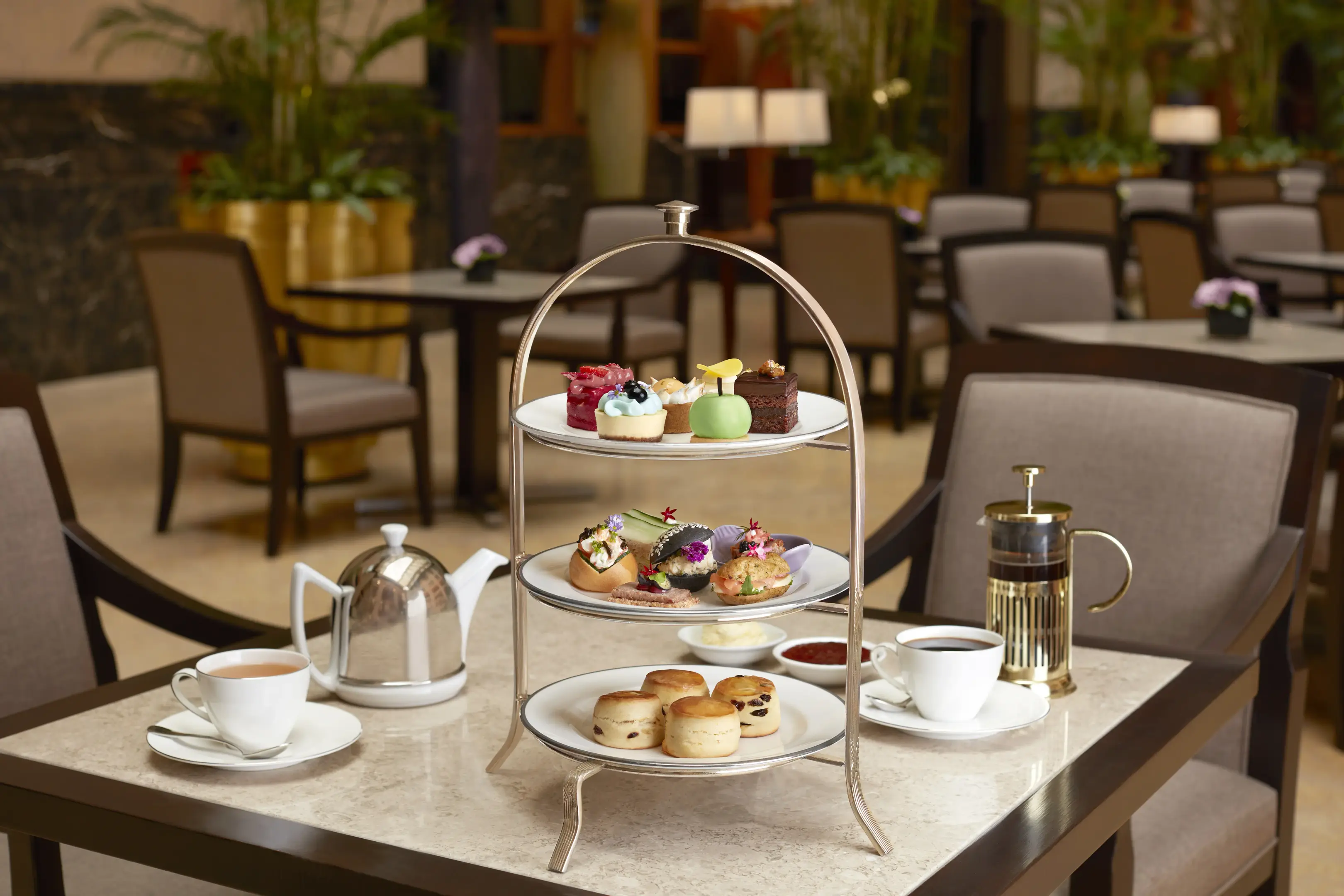 A three-tiered silver stand filled with pastries, savory sandwiches, and scones sits on a marble table arranged for afternoon tea in an elegant, sunlit courtyard lounge. Flanking the stand are a polished teapot, a French press, and two white teacups filled with warm beverages.