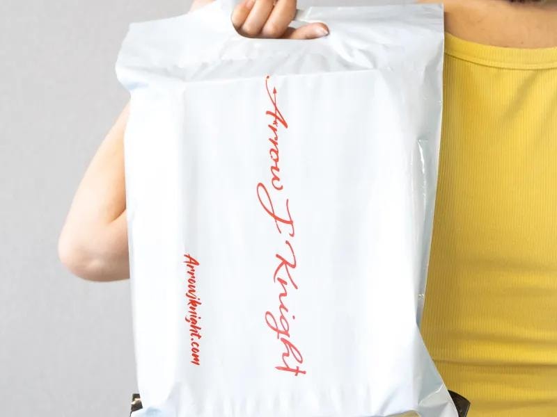 A person wearing a bright yellow t-shirt holds a branded white mailer envelope with one hand. The envelope is clearly marked with branding or a logo.