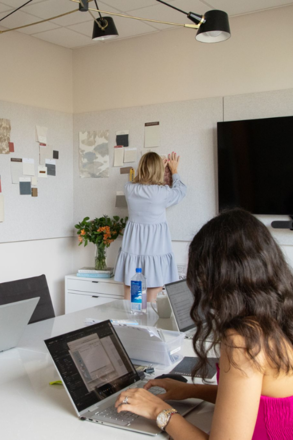 Two designers in a meeting room, one working on a laptop at a table while the other pins samples to a wall.