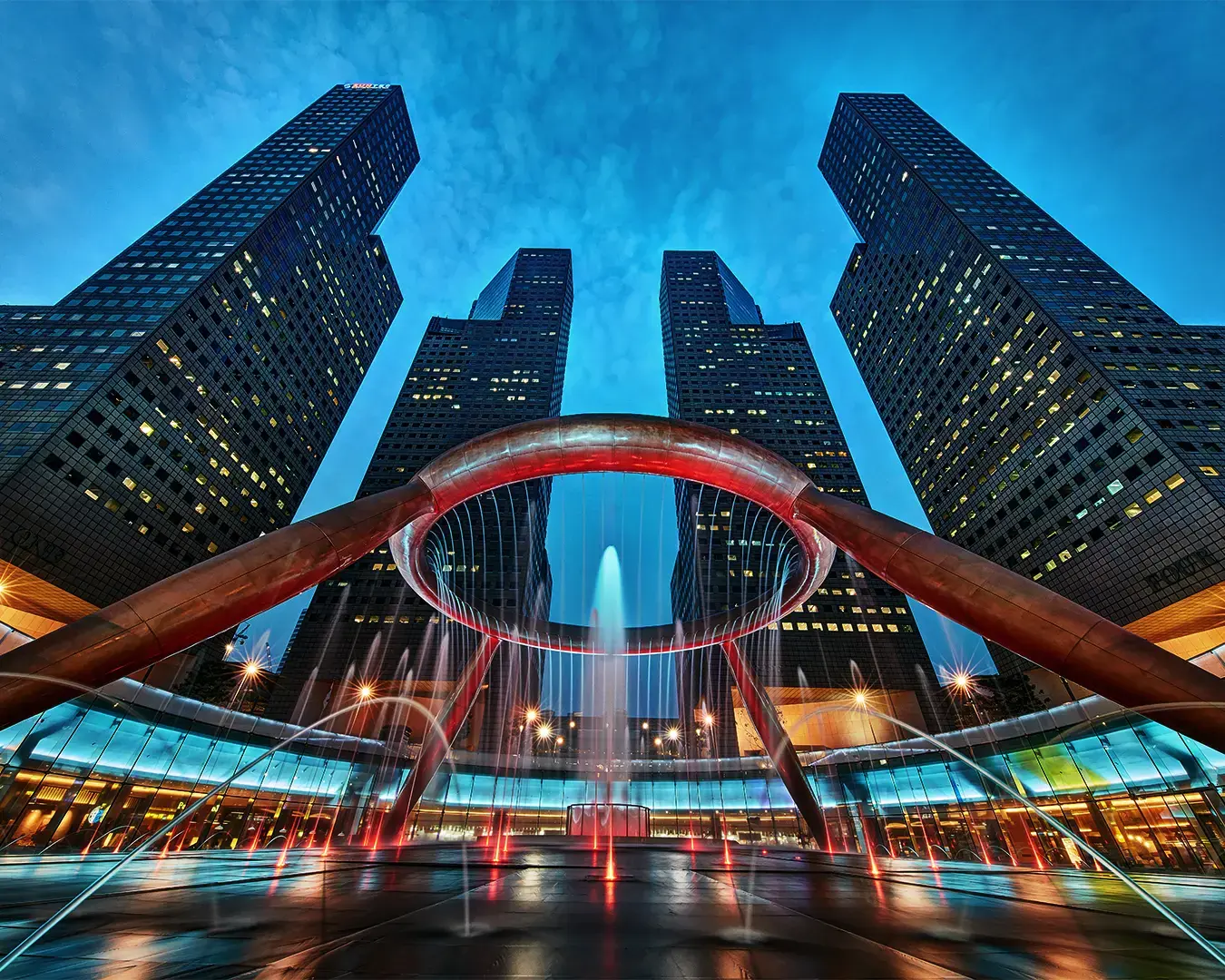 Futuristic skyscrapers with bright windows surround a large circular fountain at dusk. The sky is a deep blue, creating a dramatic urban scene.