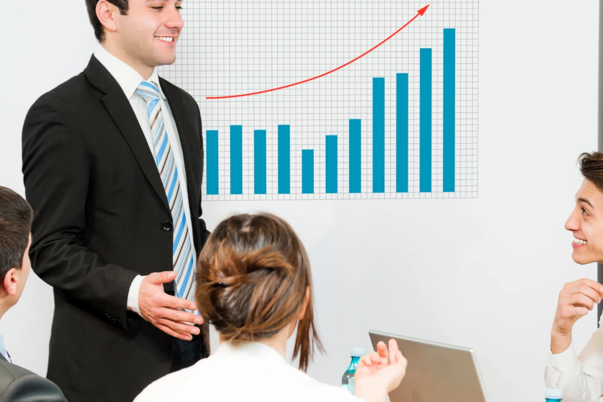 A man in a suit presents a bar graph with a rising red line to an audience in a meeting room.