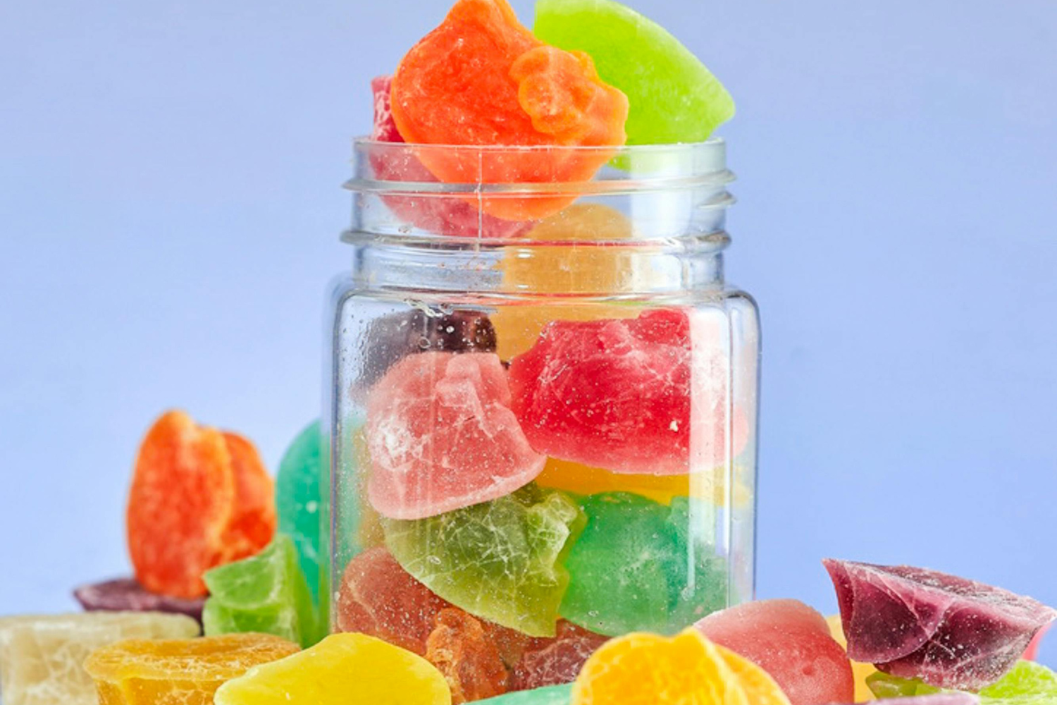 A glass jar filled with colorful gummy candies, with more scattered around the base on a light blue background.