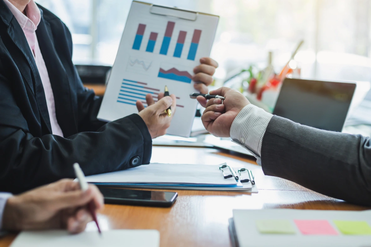 Two people in suits are looking at a clipboard with charts and graphs. One person is holding a pen, and the other is reaching for it.