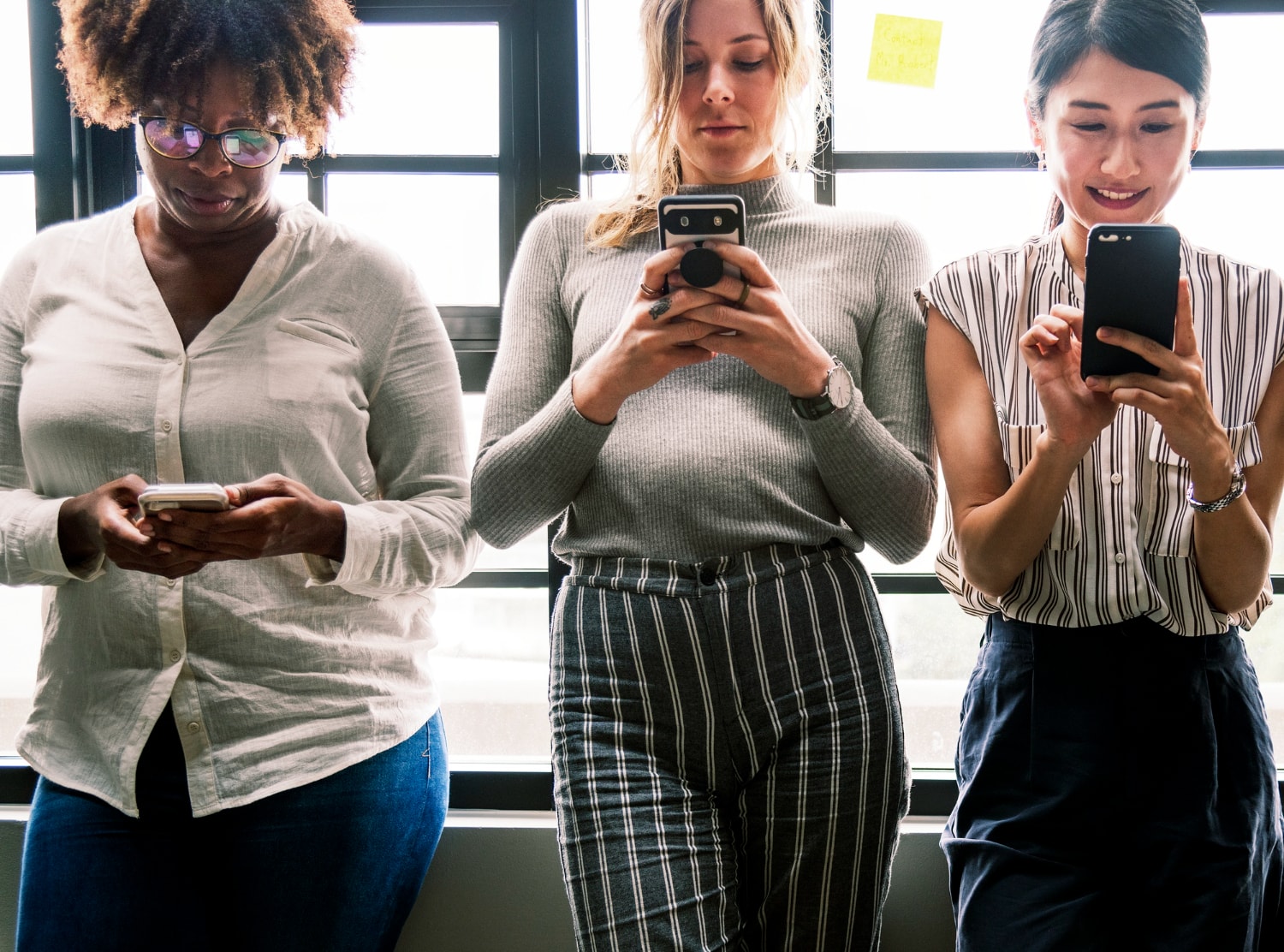 Three business professionals standing near a window, engaged with their smartphones. This represents mobile Cloud communications and Business phone solutions ensuring connectivity in a modern work environment.