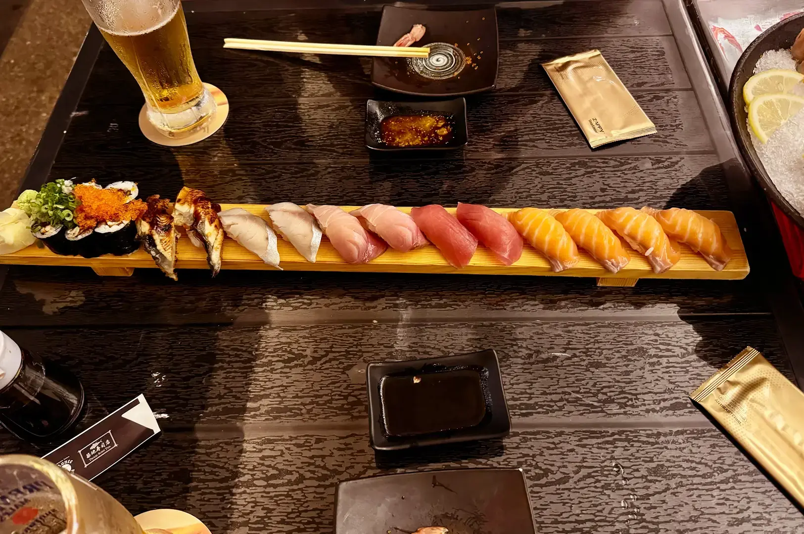 Assorted sushi and maki arranged on a wooden serving board with soy sauce, pickled ginger, wasabi, chopsticks, and a glass of beer.