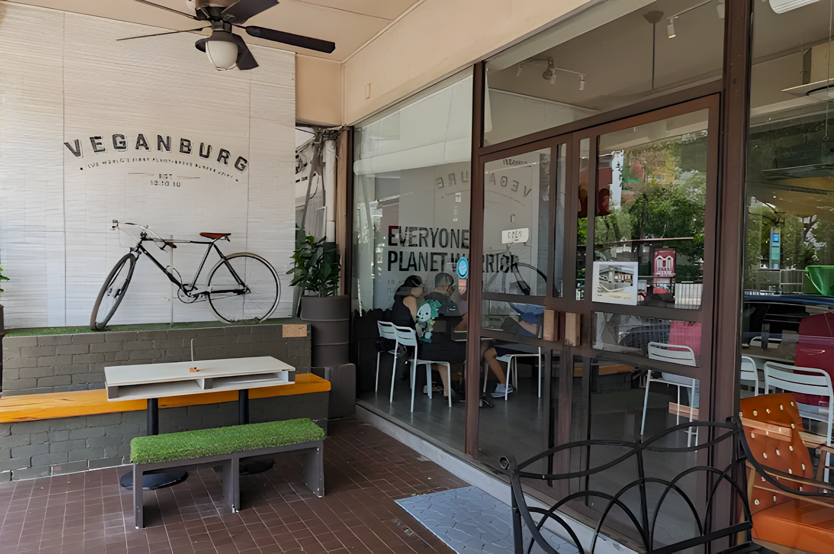 Outdoor view of a vegan restaurant with "Veganburg" signage, featuring a bicycle on the wall and seating areas. Inside, patrons enjoy meals in a casual setting.