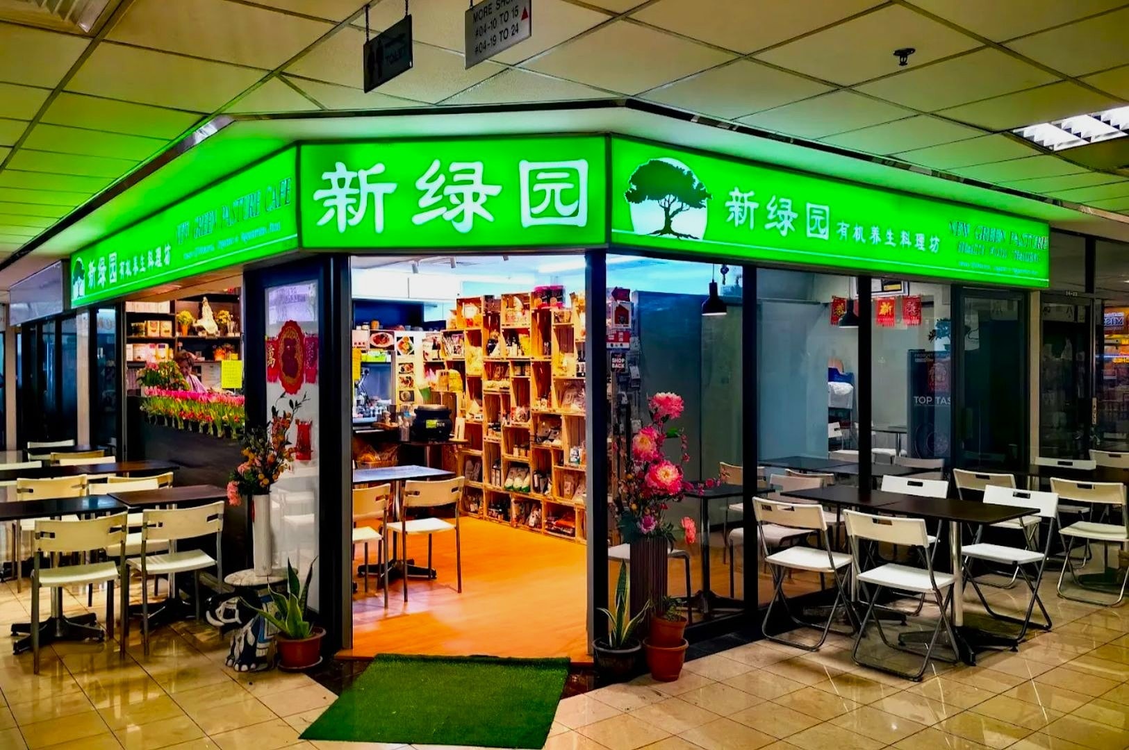 Small, brightly lit Chinese cafe with green signage and white text. Outdoor seating with white tables and chairs, flowers by the entrance, cozy ambiance.
