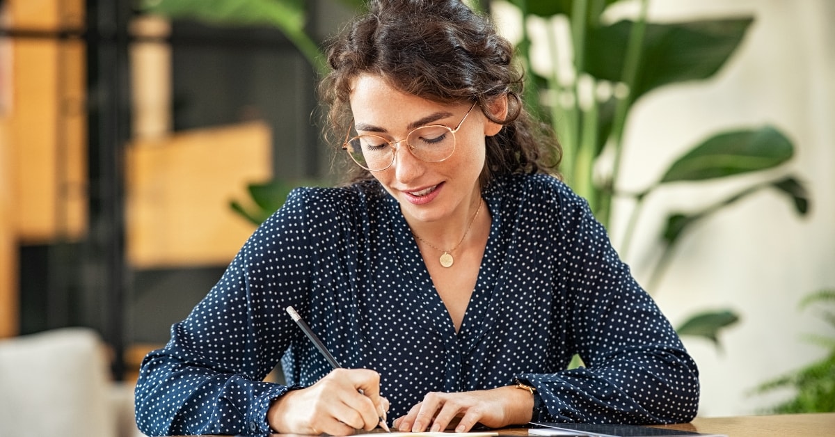 Woman with glasses writing in a notebook, organizing her OnlyFans business expenses list.