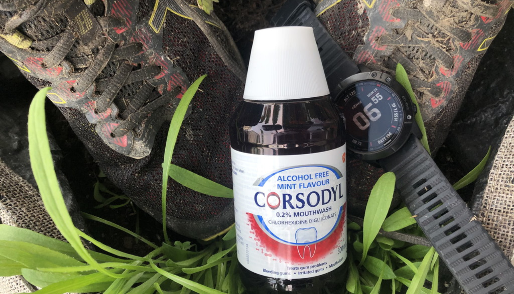 Corsodyl munnskyllflaske i naturmiljø, plassert mellom skitne joggesko og en pulsklokke, med grønt gress rundt.