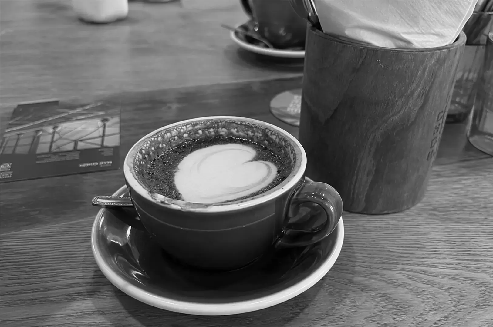 Heart latte art in a ceramic cup on a wooden café table beside a wooden utensil holder.