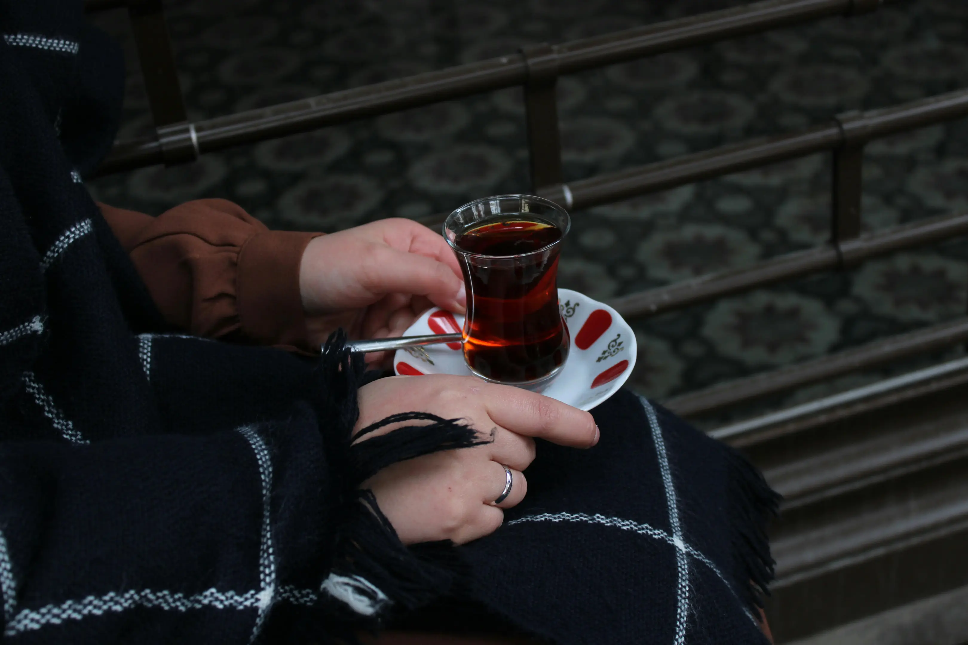A person wearing a dark plaid coat holds a traditional tulip-shaped glass of Turkish tea resting on a small patterned saucer. The person's hands are visible, with one hand wearing a silver ring, set against a dark, textured background.