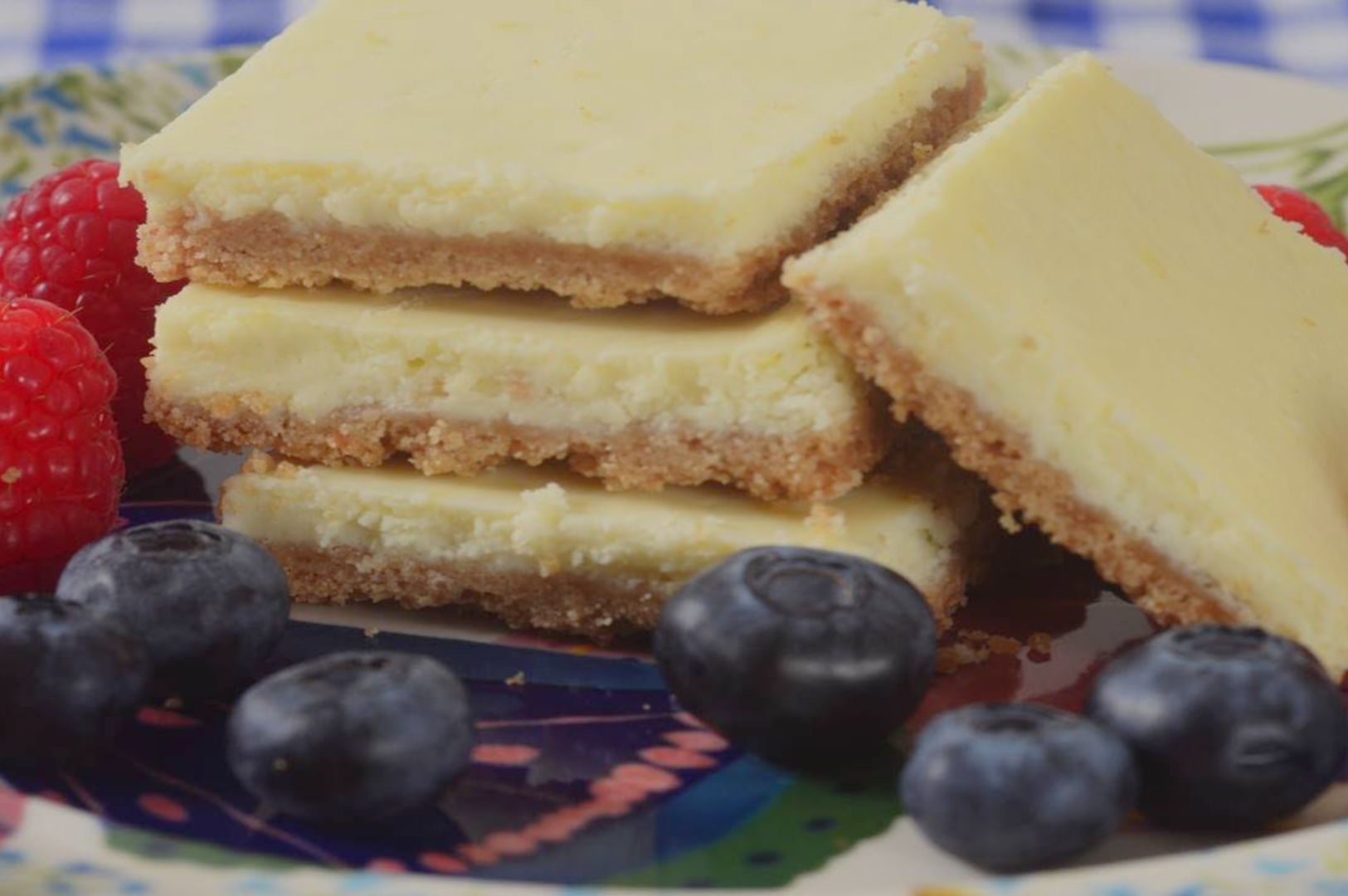 Four creamy lemon bars are stacked on a decorative plate, surrounded by fresh blueberries and raspberries, creating a vibrant and appetizing scene.