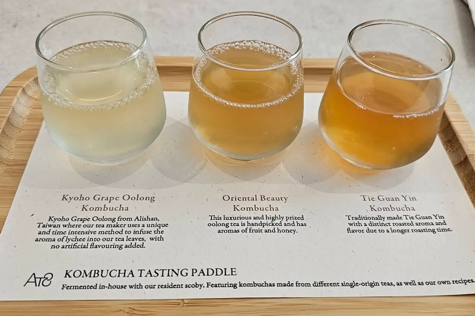 A top-down shot focuses on a "Kombucha Tasting Paddle" set on a light wooden tray over a printed description sheet. Three stemless glass tumblers are arranged in a row, each containing a different shade of kombucha ranging from a pale golden-yellow "Kyoho Grape Oolong" on the left, to a warm amber "Oriental Beauty" in the center, and a deeper reddish-brown "Tie Guan Yin" on the right, all showing delicate effervescent bubbles at the surface.