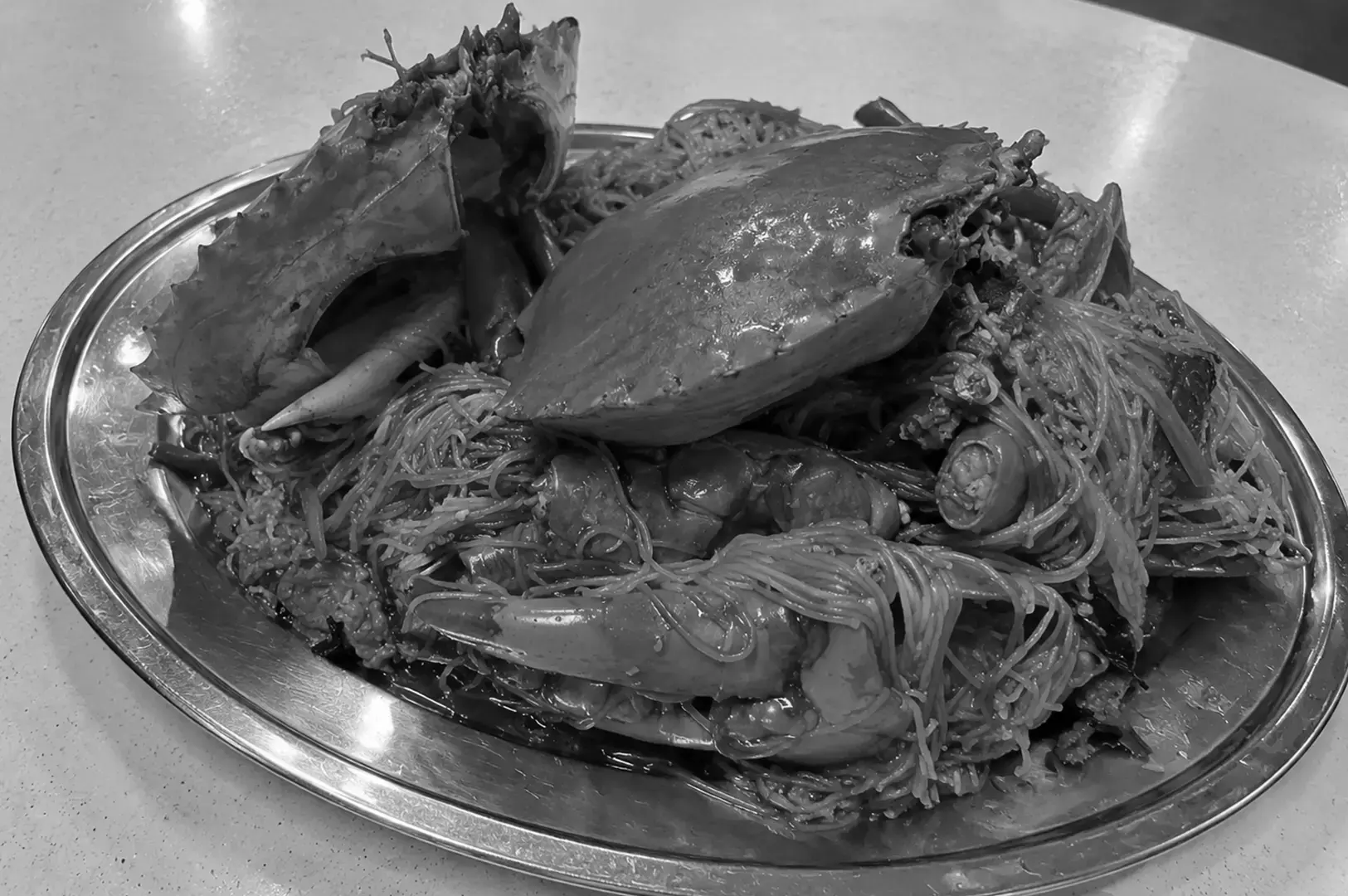 A large seafood platter with whole crab, oversized claws, and noodles coated in sauce, served on a shiny oval metal tray on a light-colored table.