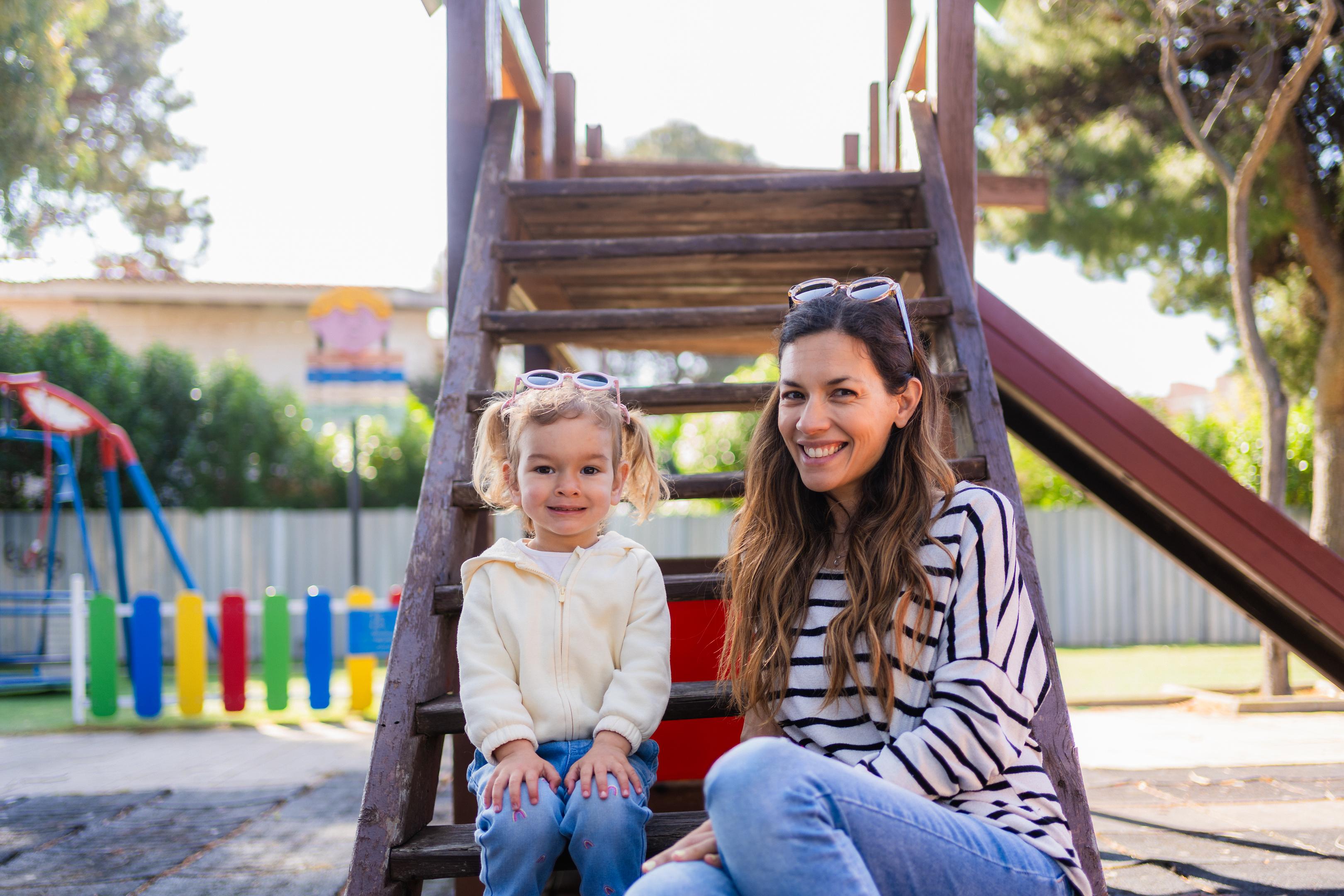 A young girl and a woman are sitting on the steps of a playground structure on a sunny day.
