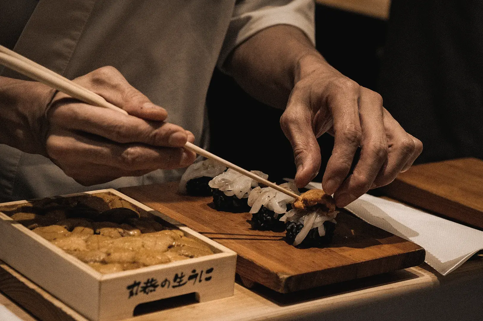 A medium close-up action shot shows the sushi chef using chopsticks to carefully place fresh sea urchin onto sushi at Sushi to Sumi omakase counter.