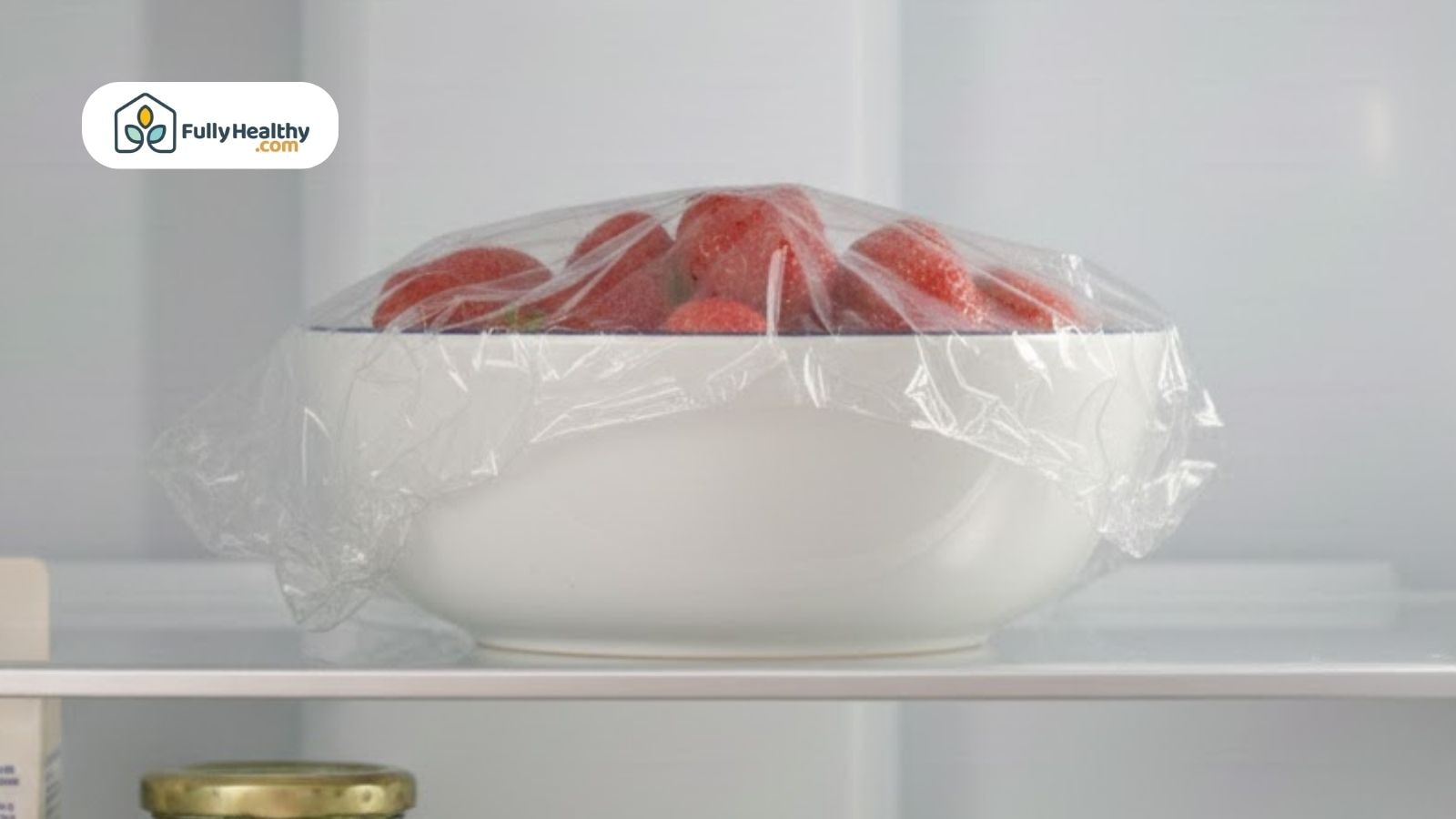 Strawberries covered with plastic wrap in shallow bowl inside refrigerator.