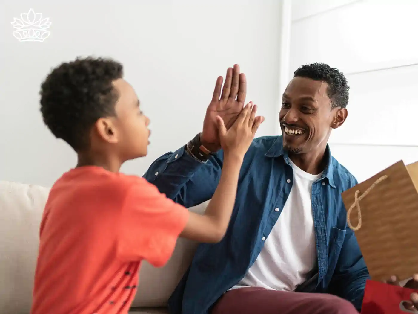 A joyful father and his young son high-fiving on a sofa, celebrating a moment of happiness together. The father holds a gift bag, suggesting a special occasion. 