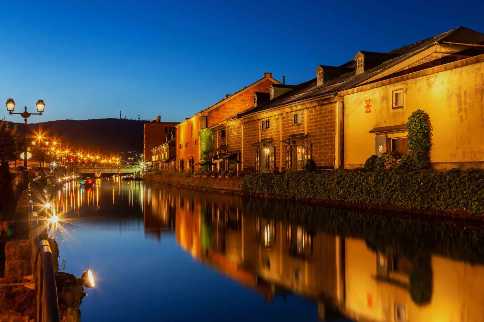 A serene canal scene at dusk, featuring illuminated buildings along the water's edge, with reflections creating a symmetrical view.