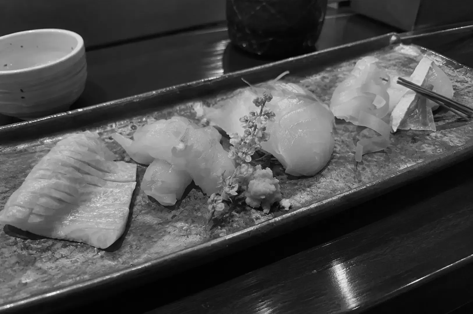 Assortment of sliced sashimi arranged across a long ceramic platter with flowers, garnishes, and a small bowl of wasabi.