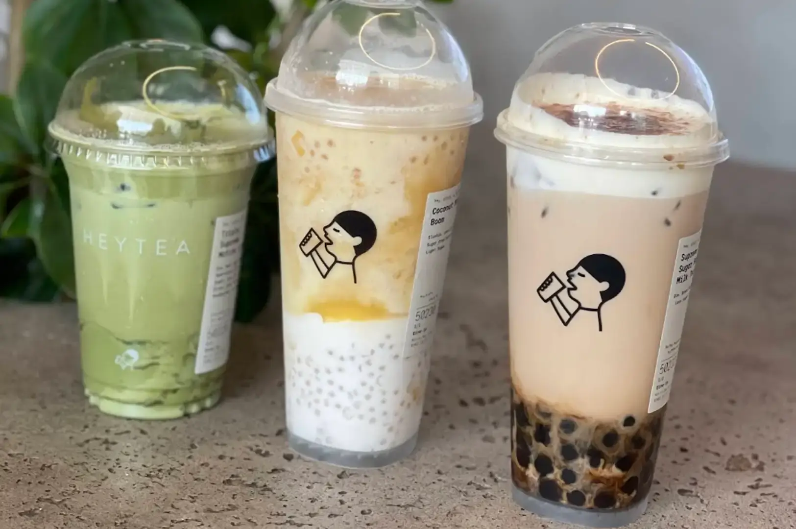 Three distinct HEYTEA beverages in clear plastic cups featuring the iconic drinking-person logo are lined up on a counter: a vibrant green iced matcha latte on the left, a layered coconut mango sago smoothie in the middle, and a brown sugar boba milk tea with cheese foam on the right.