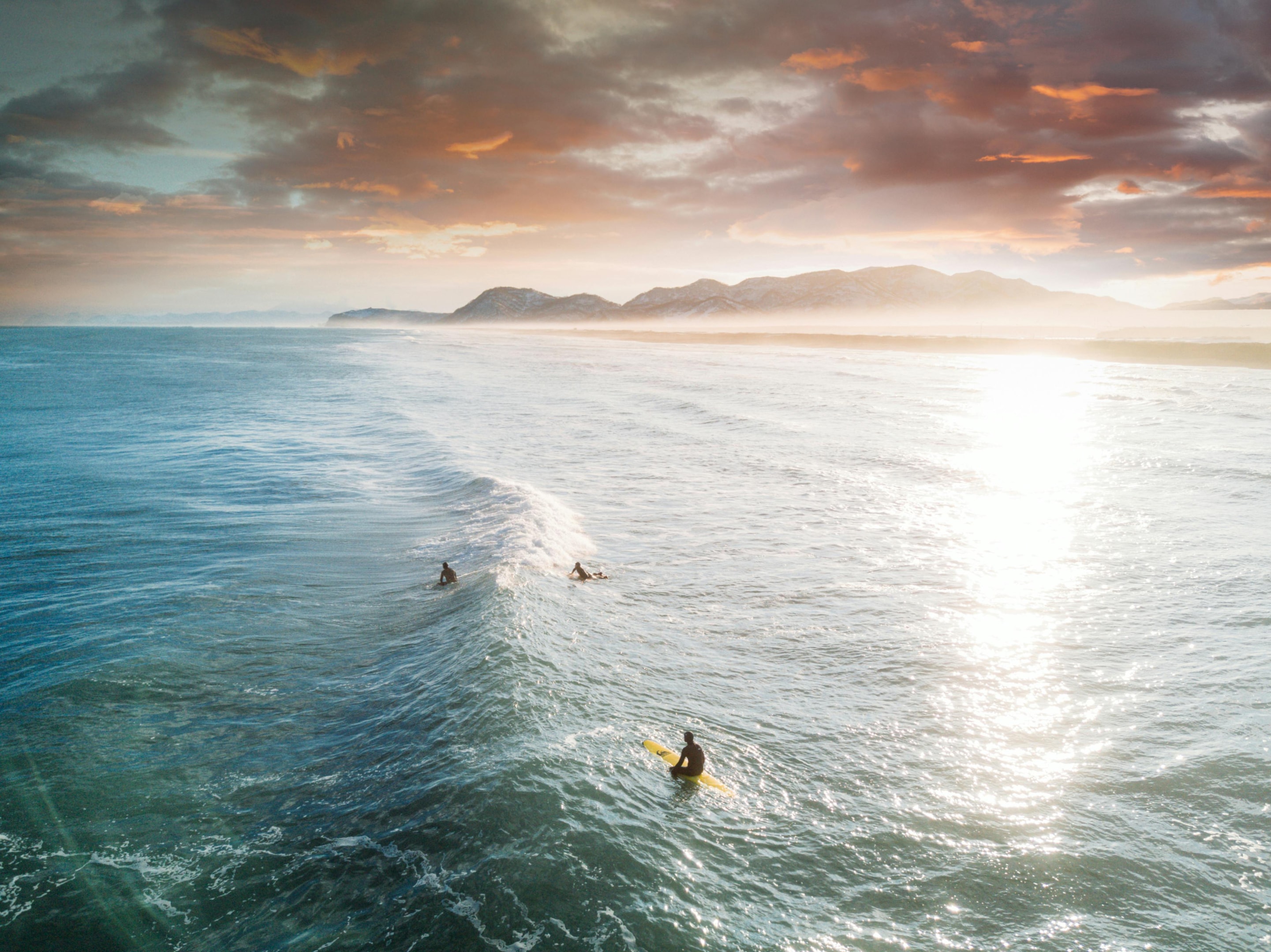 Photo de Yaroslav Shuraev. Photograph of surfers paddling out on the waves at sunset, with the sun casting a golden glow over the ocean and distant mountains in the background.