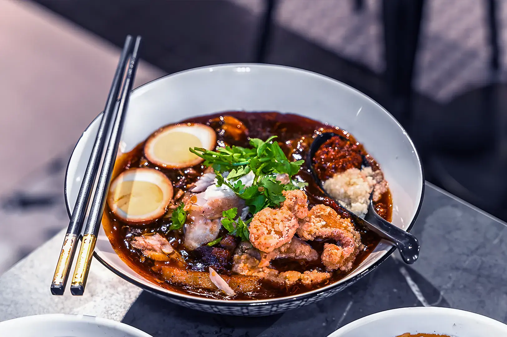 A flavorful bowl of ramen with rich broth, topped with crispy chicken, soft-boiled eggs, cilantro, and spices. Gold-tipped chopsticks rest on the bowl's edge.
