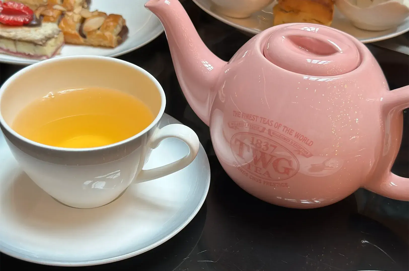 A close-up shot of a classic light pink TWG ceramic teapot and a filled teacup served with fresh pastries during an afternoon high tea.