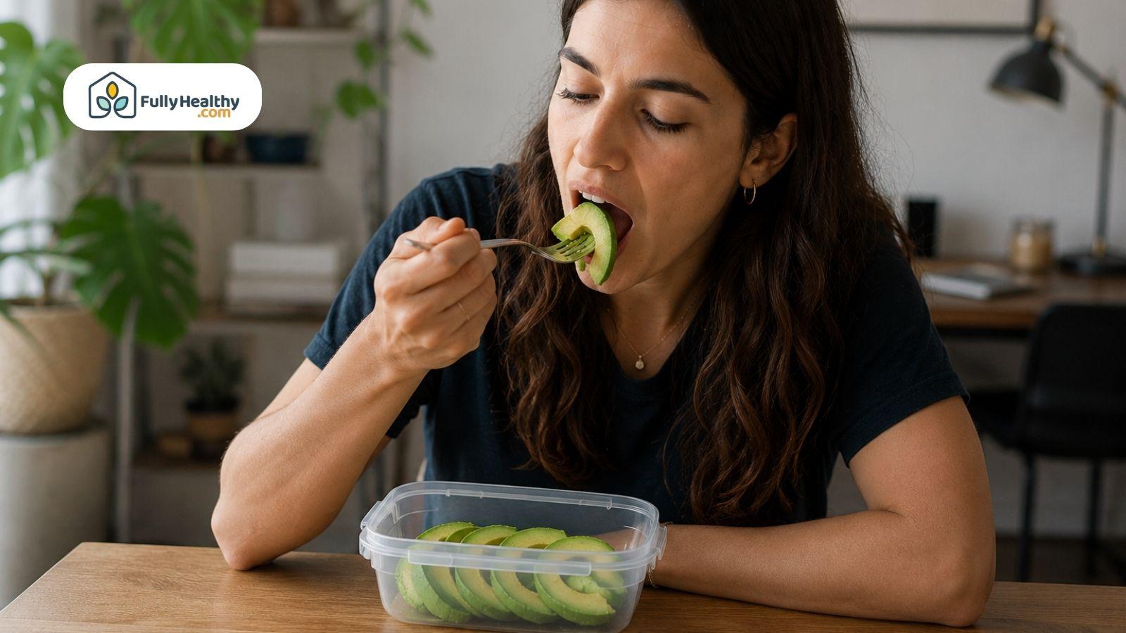 Woman eating fresh avocado slices from a container at home kitchen table.