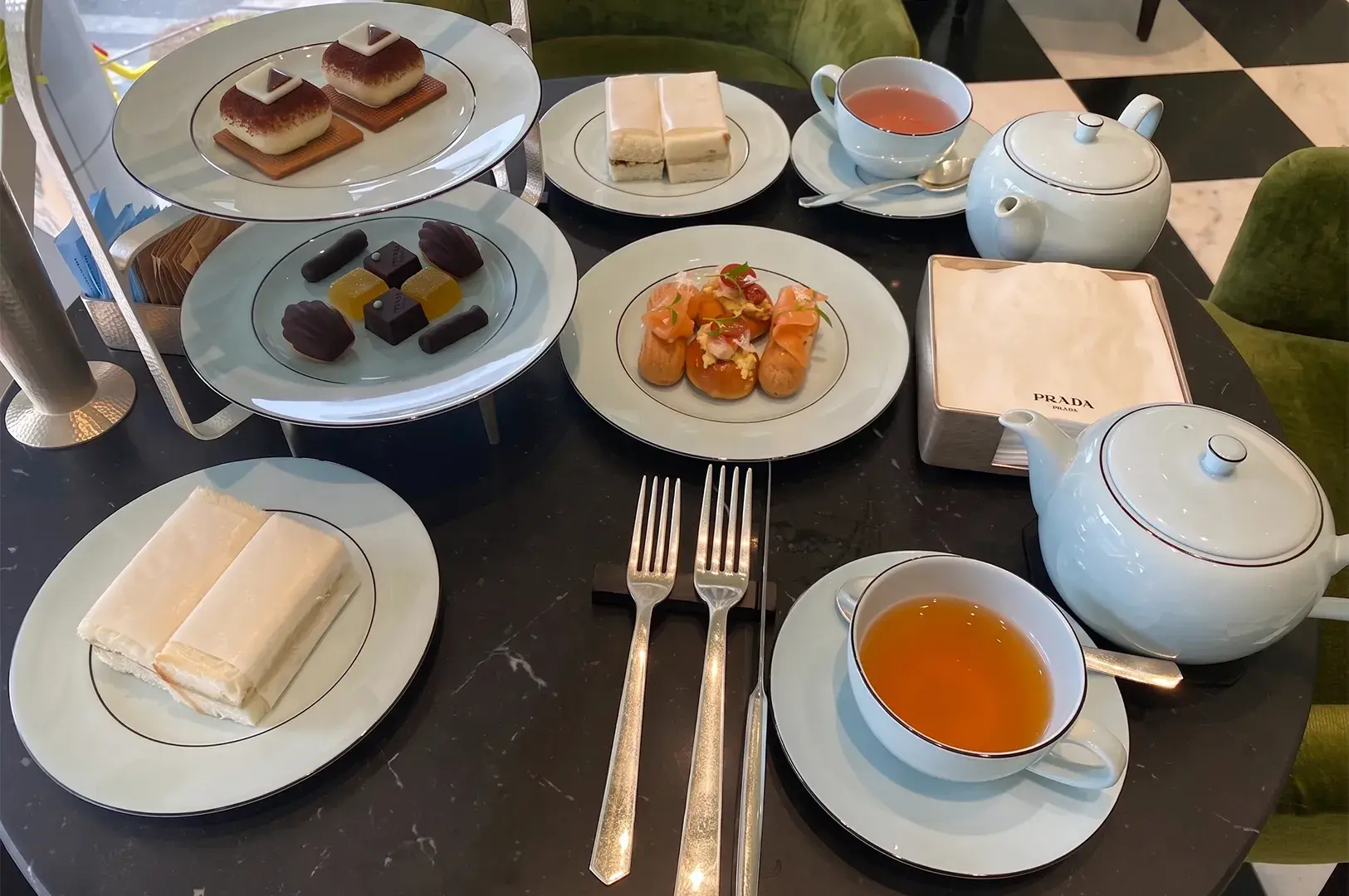 Elegant afternoon tea table set with mint-green porcelain teaware, tea sandwiches, fine pastries, chocolates, savory bites, and a Prada-branded napkin box.