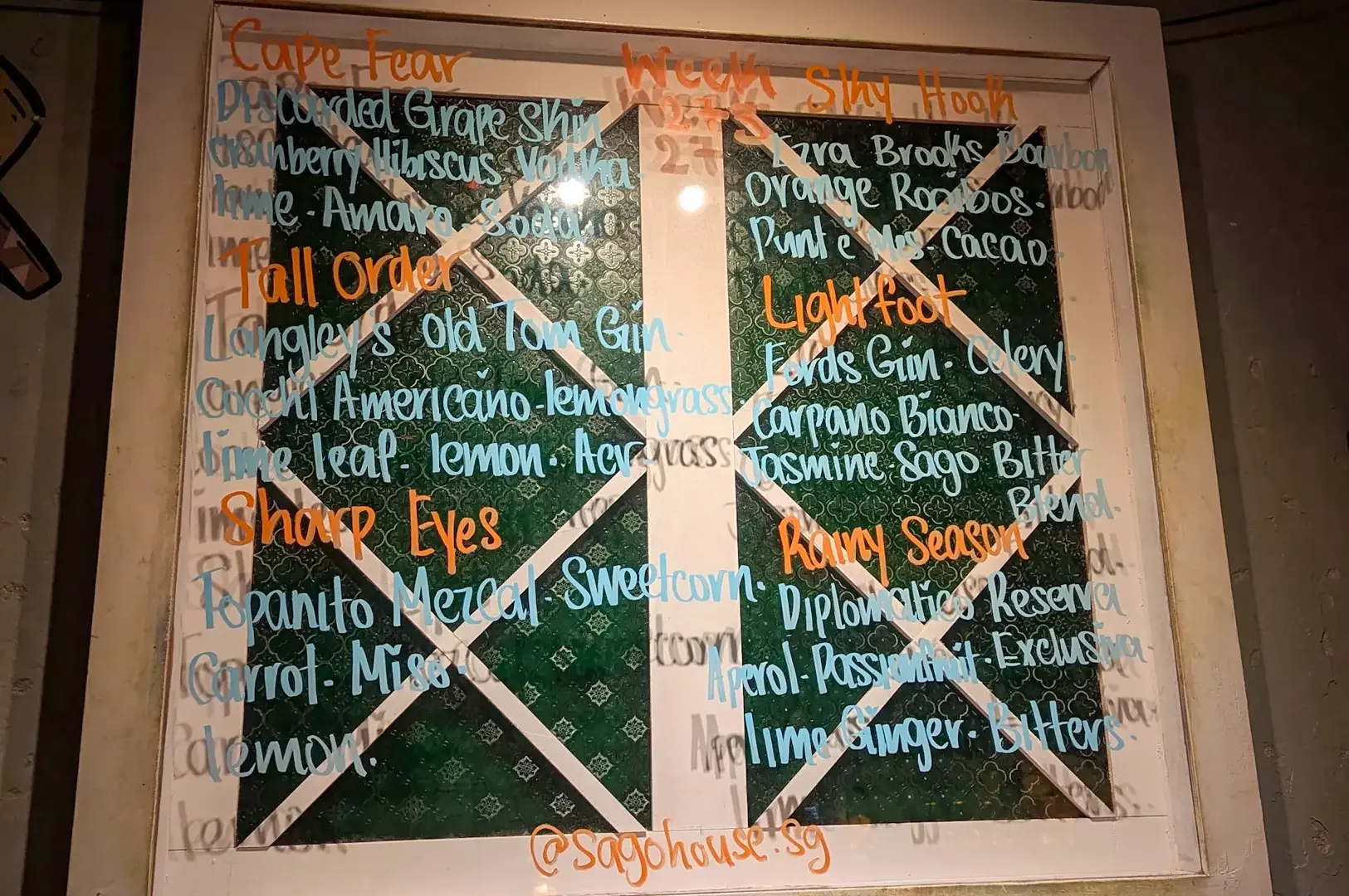 Straight-on eye-level shot of a handwritten cocktail menu board displayed indoors at a bar, featuring multiple drink names, ingredients, and section headings written in colorful chalk markers.