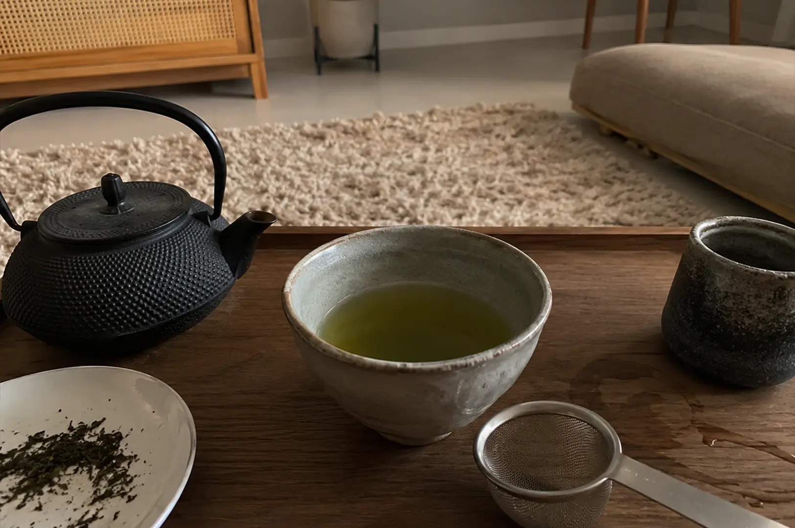 A close-up, high-angle shot features a tea preparation set arranged on a dark wooden tray. In the center sits a rustic, off-white ceramic bowl filled with green tea. To its left is a black, textured cast-iron teapot with a curved handle, and below it, a small white plate holding loose tea leaves. To the right of the main bowl is a taller, dark textured ceramic cup, and in the lower right corner, a metal mesh tea strainer rests on the wood. The background shows a cozy room interior, with a fuzzy cream-colored rug and a beige cushion or sofa out of focus.