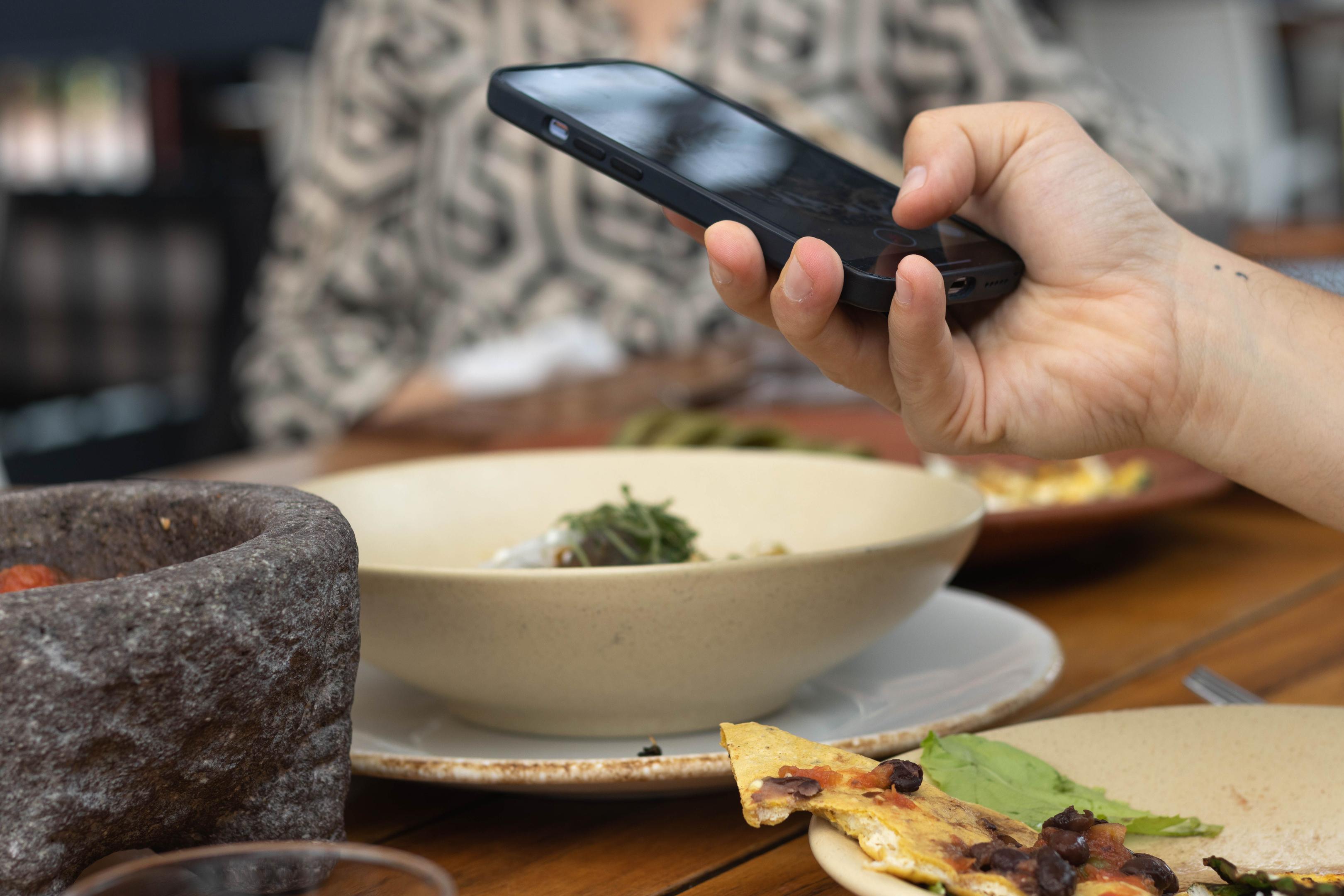 A person is sitting at a cafe, intently scrolling through a restaurant menu on their smartphone, exploring various menu items to enhance their dining experience. This digital engagement reflects the importance of having a mobile-friendly restaurant website to attract new customers and build loyalty among existing diners.