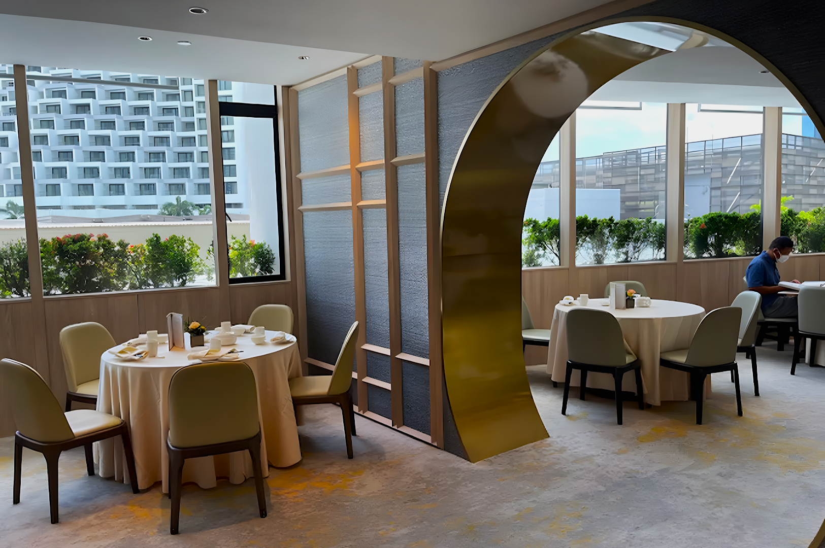 Modern dining room with round tables, cream chairs, and elegant decor. Large windows offer a view of greenery and an adjacent building, creating a serene ambiance.
