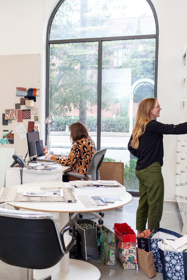 Interior design studio workspace with team members organizing samples and working at desks near a large arched window.