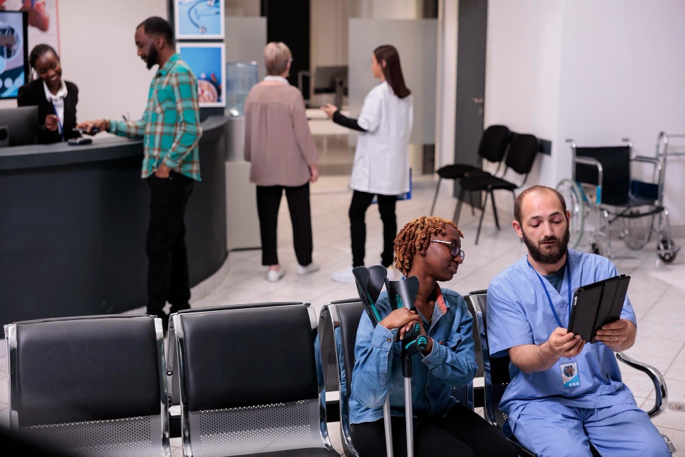 A nurse in scrubs holds a tablet and consults with a seated patient on crutches in a busy hospital waiting area, while other staff and patients interact at a nearby reception. Unified Communications and team collaboration tools support seamless communication and streamline business productivity by enhancing patient intake and triage processes.