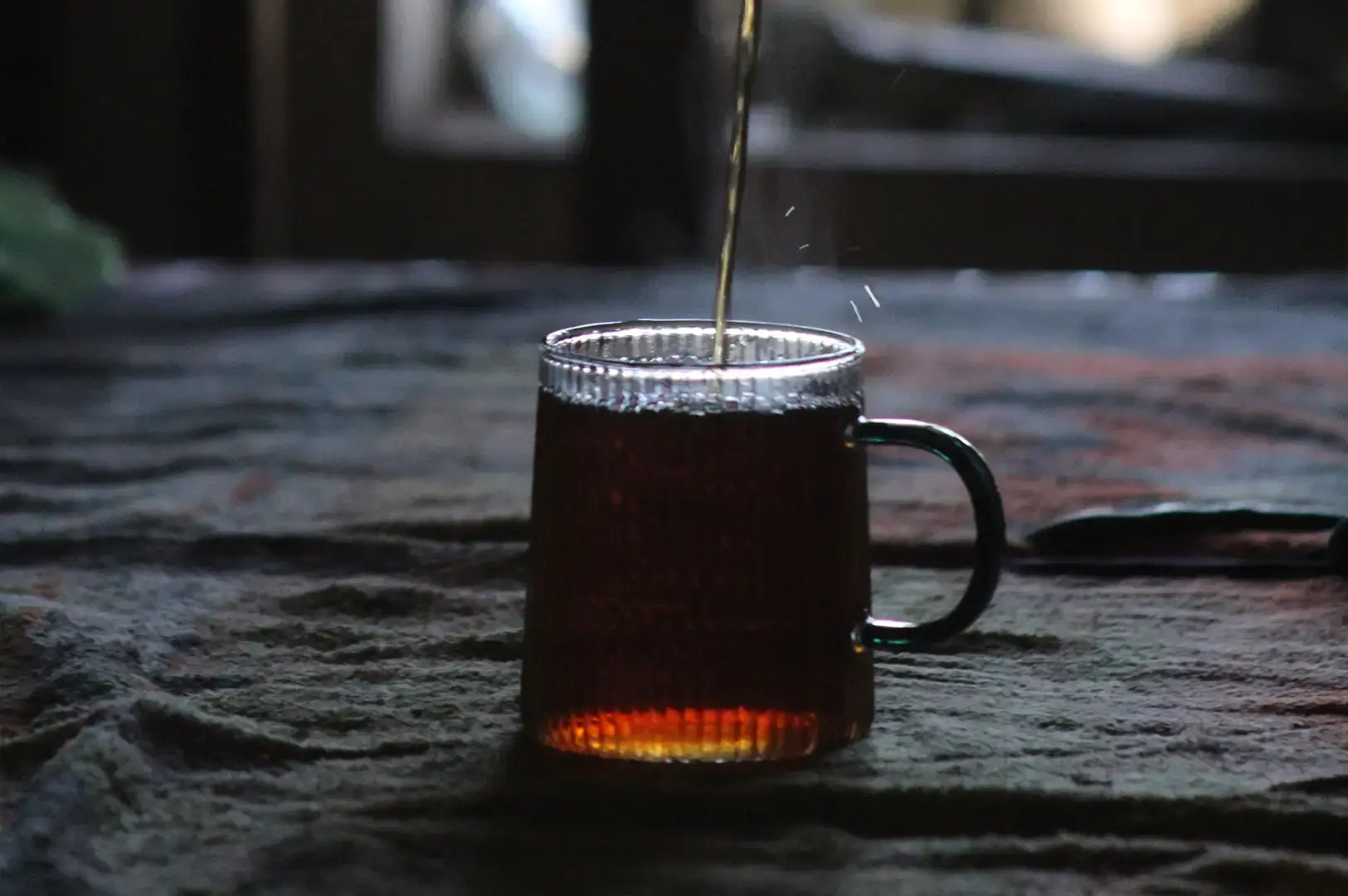 A stream of freshly brewed tea pours into a clear, ribbed glass mug sitting on a textured dark fabric tablecloth in a moody, low-light setting.