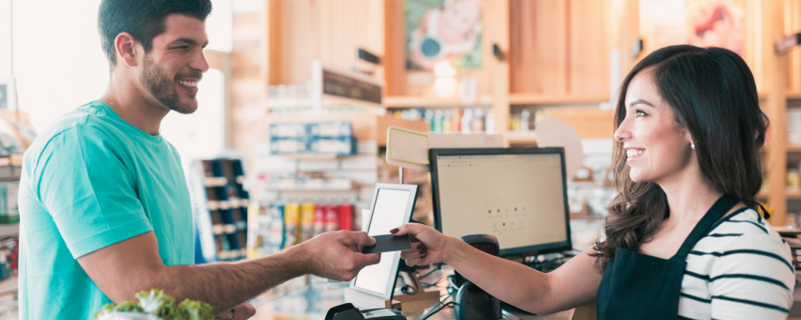 Imagem: Uma caixa registradora em um balcão de loja com um cliente entregando dinheiro ao vendedor. ALT: "Vendas Transacionais: Cliente pagando em dinheiro no balcão de uma loja