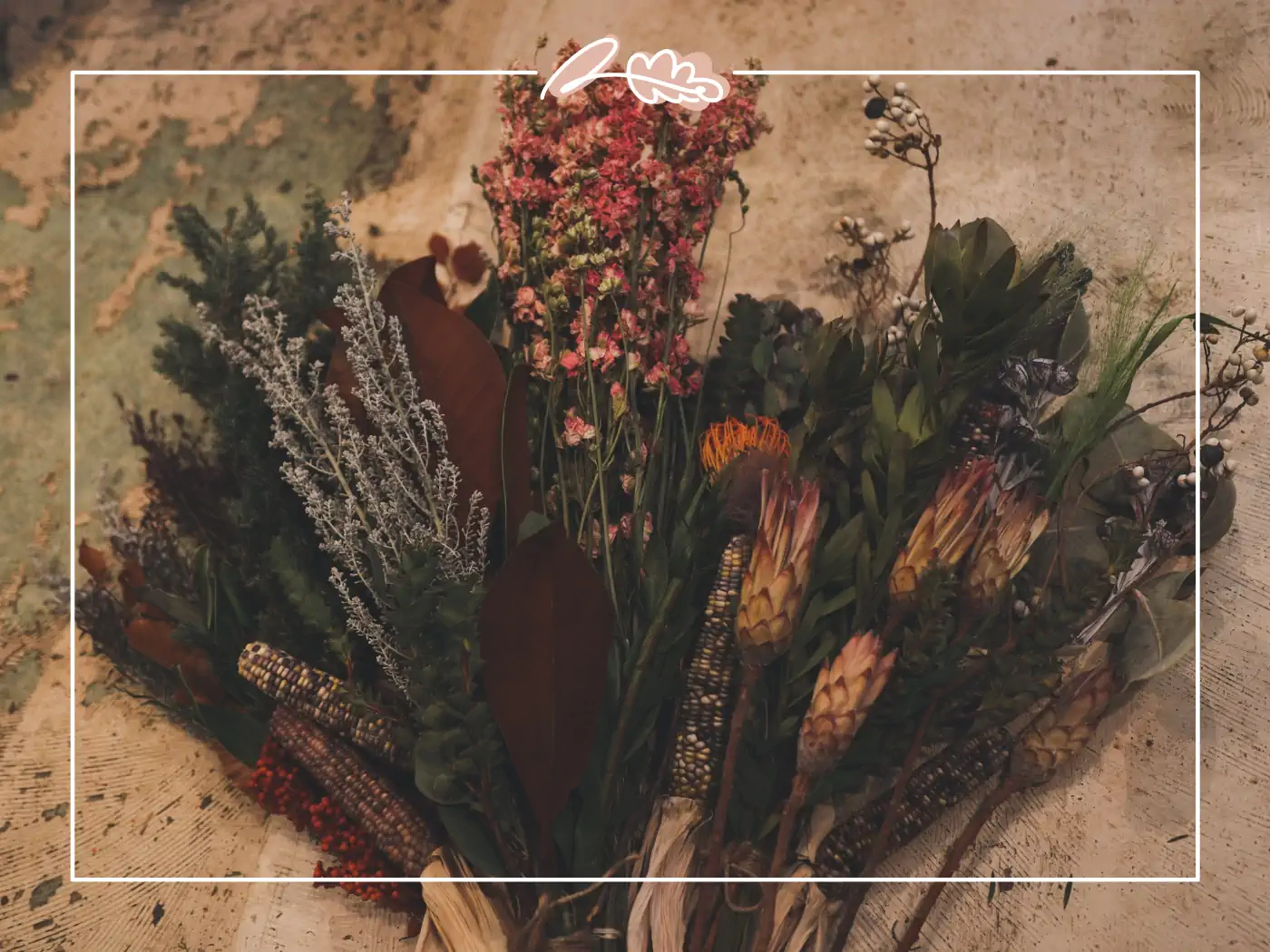 A beautifully arranged rustic bouquet of dried flowers featuring a mix of green foliage, pink blossoms, and other dried elements, lying on a textured background. Fabulous Flowers and Gifts.