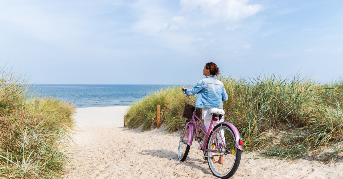 Woman walking a lavender beach cruiser bicycle down a sandy dune path toward the ocean under a partly cloudy sky.