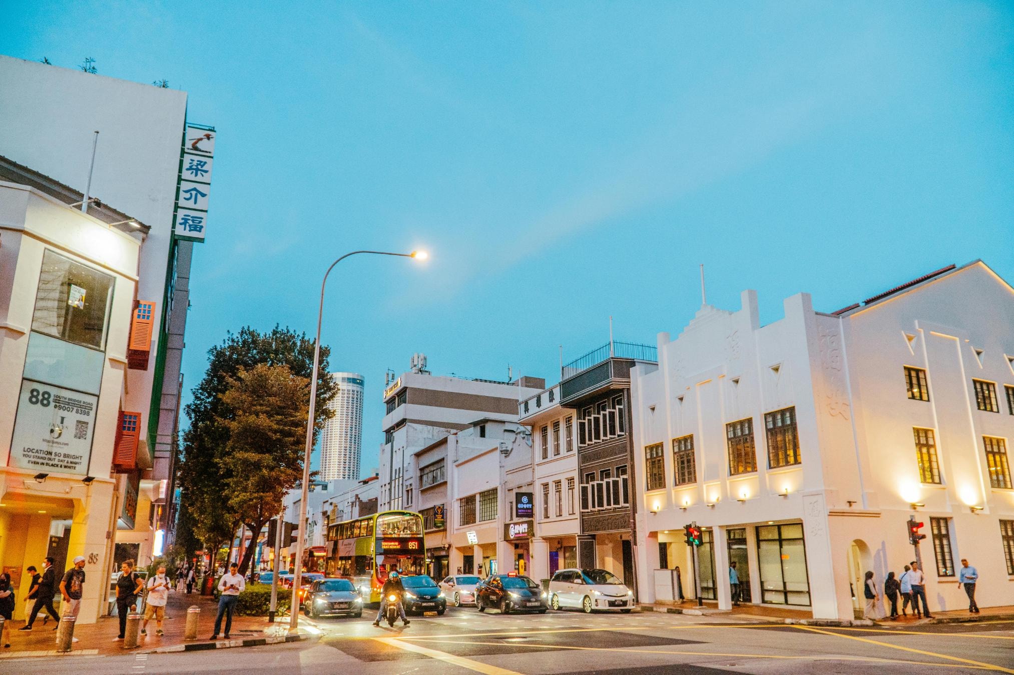 Illuminated white heritage buildings line a busy street intersection at twilight, casting a warm glow on the scene. Pedestrians stroll along the sidewalks while a double-decker bus and cars wait at the traffic lights under a deepening blue sky.