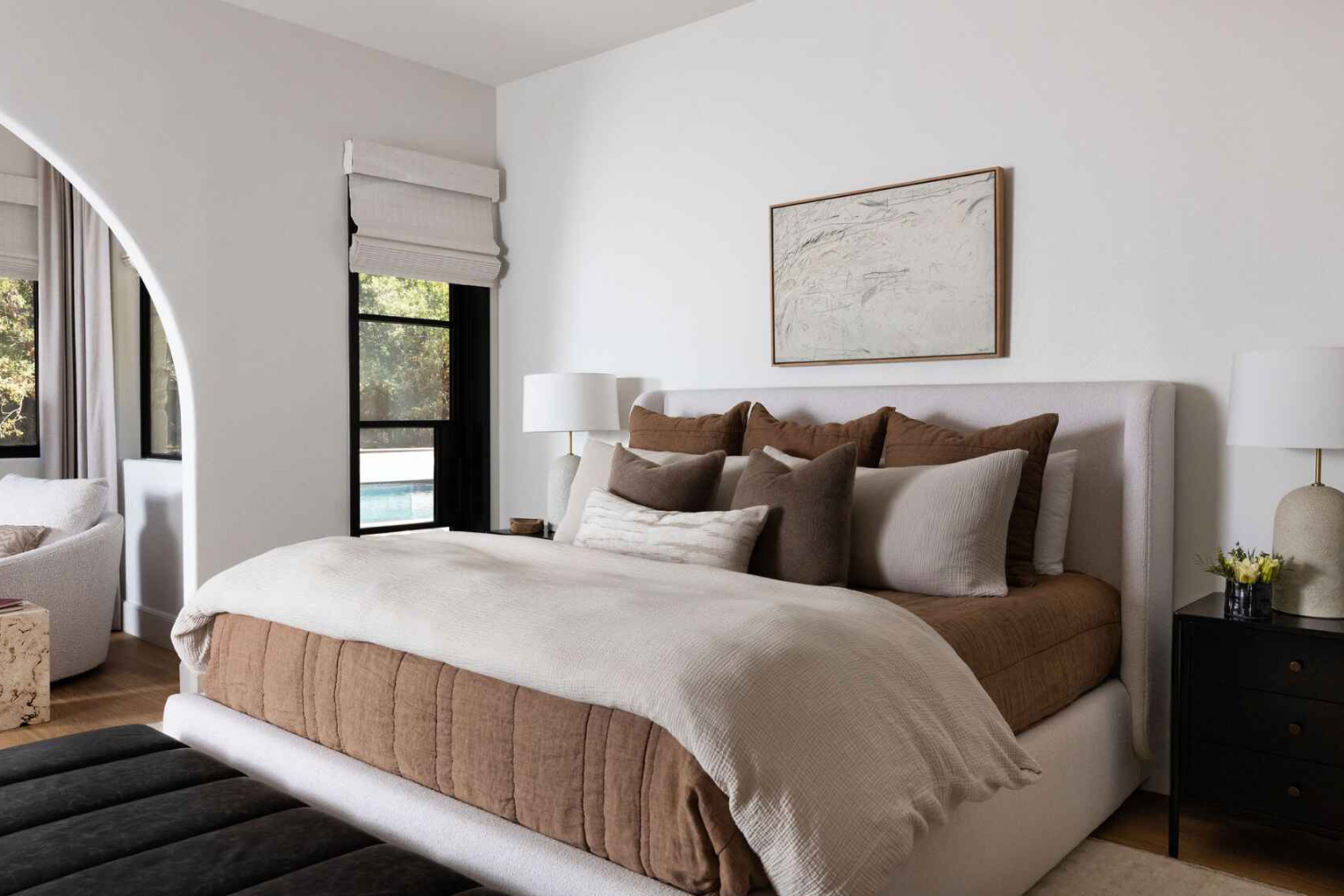 Neutral-toned bedroom with a soft upholstered bed, brown and cream bedding, arched doorway, and black window trim framing an outdoor pool vi