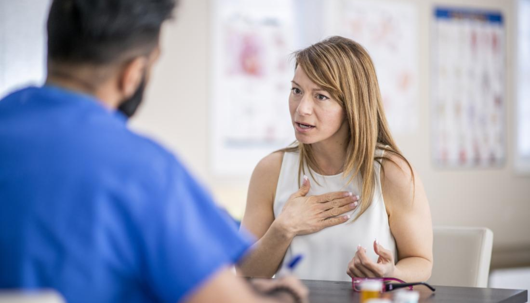 Kvinne forklarer symptomer og holder hånden på brystet mens hun snakker med lege i blått antrekk på et legekontor.