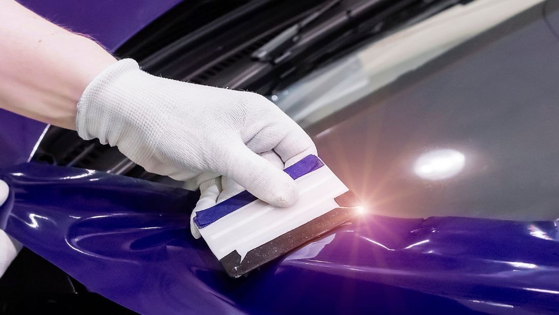 Close-up of a gloved hand using a squeegee to smooth out a purple vinyl wrap on a car’s hood, showing the precision required in professional cargo van wrap design.